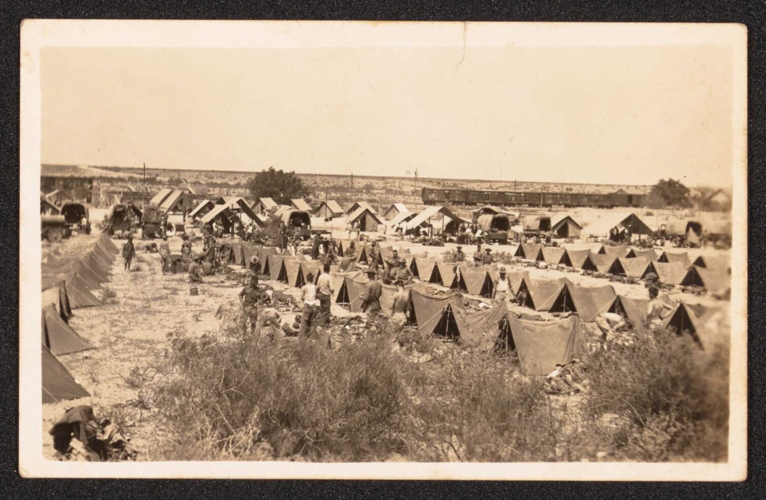 A large outdoor military camp with rows of tents and people working. Trucks are parked nearby.