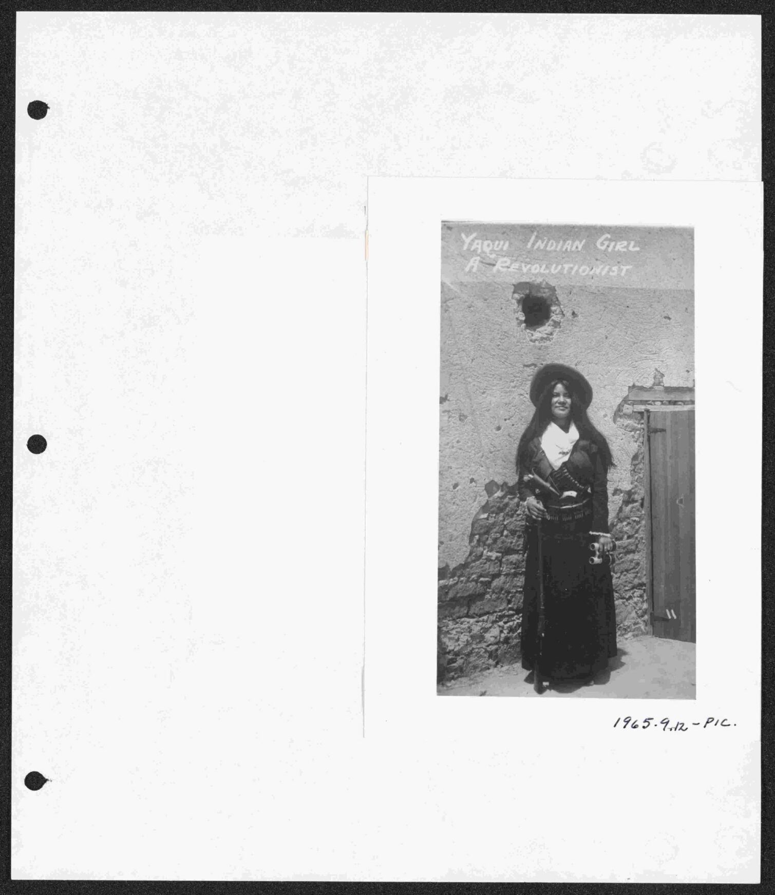 A young Yaqui Indian girl in traditional dress with a hat, standing against a wall.