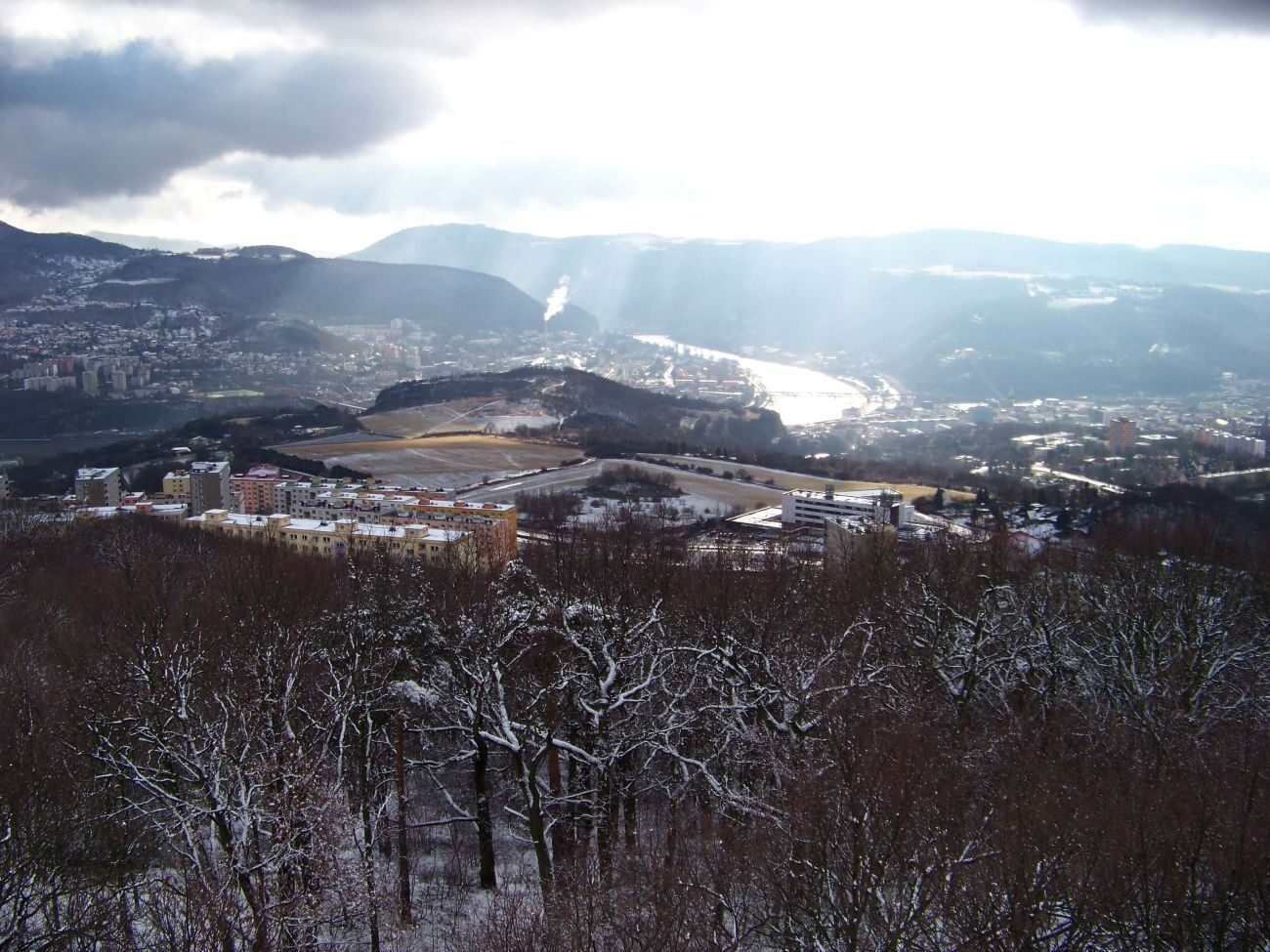 A snowy landscape viewed from a hill, showing a small town and mountains in the distance.